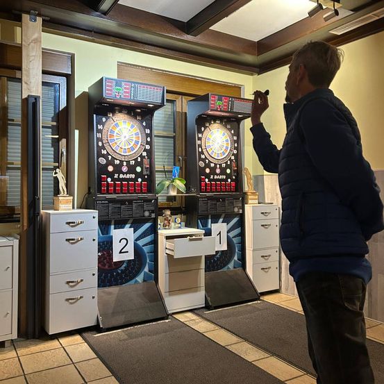 Ein Mann steht vor zwei Dart-Automaten, der eine mit '2' und der andere mit '1' beschriftet. Er hält einen Dart und zielt auf das Brett.