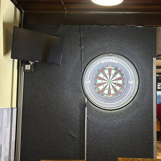 Ein Indoor-Dartboard mit dem Wort Winmau, montiert an einer Wand mit einem Bildschirm. Zwei Personen sitzen auf Stühlen an einem Tisch, während andere vor der Theke stehen.