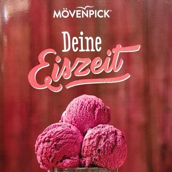 Eine Nahaufnahme eines Desserts, wahrscheinlich rosa Eis, in einer metallischen Schüssel. Die Schüssel steht auf einem Tisch mit einem roten Tuch. Dahinter befinden sich Brombeeren. Der Hintergrund ist unscharf.