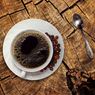 Eine Tasse schwarzen Kaffees steht auf einer Untertasse mit Kaffeebohnen auf einer Holzoberfläche. Ein silberner Löffel ist rechts platziert.