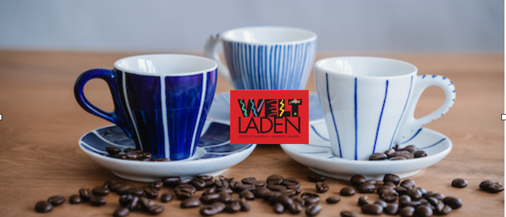 Drei Kaffeetassen mit blauen Streifen stehen auf Untertassen mit verstreuten Kaffeebohnen auf einem Holztisch. Die Tassen sind blau, weiß und blau-weiß. Ein rotes Logo mit dem Text 'Welt Laden' ist sichtbar.