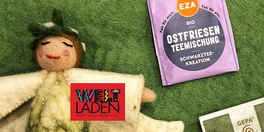 Eine Puppe in Weiß liegt auf einer grünen Oberfläche neben einer lila Tasche mit der Aufschrift Ostfriesen Teemischung, einem roten Logo und einem Stück Papier.
