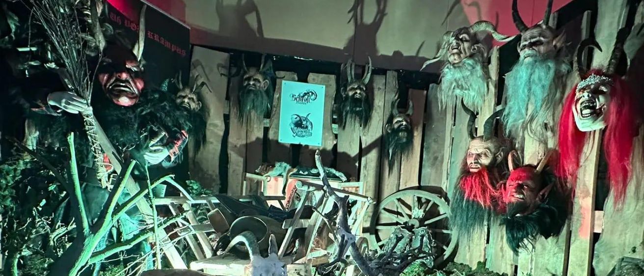 Ein dunkler Raum mit Holzwänden zeigt zahlreiche Hörner, ein Schild und verschiedene Masken. Ein Schild steht 'Hexennacht'. Fahrräder und andere Gegenstände sind verstreut.