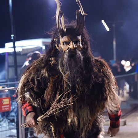 Eine Person in einem Kostüm mit Hörnern und Fell geht bei Nacht. Ein Publikum beobachtet im Hintergrund.