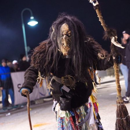 Eine Person in einem gruseligen Kostüm mit Maske, Stock und Besen geht im Dunkeln. Dahinter steht eine Menschenmenge, eine Straßenlampe und ein Banner.