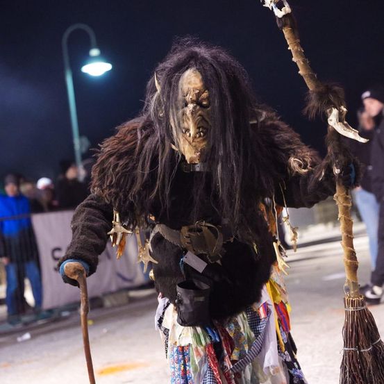 Eine Person in einem gruseligen Kostüm mit Maske, Stock und Besen geht im Dunkeln. Dahinter steht eine Menschenmenge, eine Straßenlampe und ein Banner.