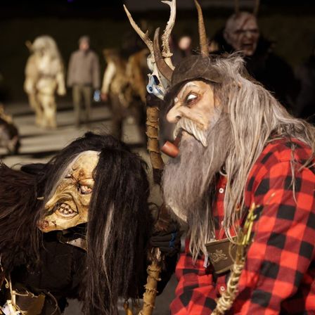 Zwei kostümierte Personen mit Masken und Hörnern auf den Köpfen gehen zusammen. Die Person auf der linken Seite trägt ein schwarzes Outfit mit langen Haaren. Die Person auf der rechten Seite trägt ein rot-schwarz kariertes Hemd.