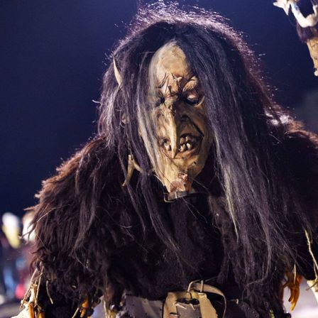 Eine Person in einem monströsen Kostüm mit langen, wilden Haaren und einem furchterregenden Gesicht. Das Kostüm hat eine raue Fell, scharfe Hörner und einen Gürtel. Die Person scheint bei einer Halloween-Veranstaltung aufzutreten.