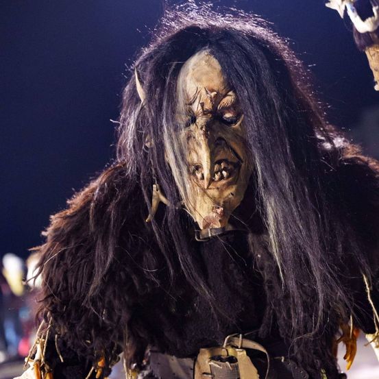 Eine Person in einem monströsen Kostüm mit langen, wilden Haaren und einem furchterregenden Gesicht. Das Kostüm hat eine raue Fell, scharfe Hörner und einen Gürtel. Die Person scheint bei einer Halloween-Veranstaltung aufzutreten.