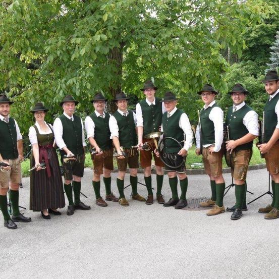 Eine Gruppe von Menschen in traditioneller bayerischer Kleidung, einschließlich grüner Westen und Hüten, steht im Freien zusammen und hält Musikinstrumente. Bäume und Grünfläche im Hintergrund.