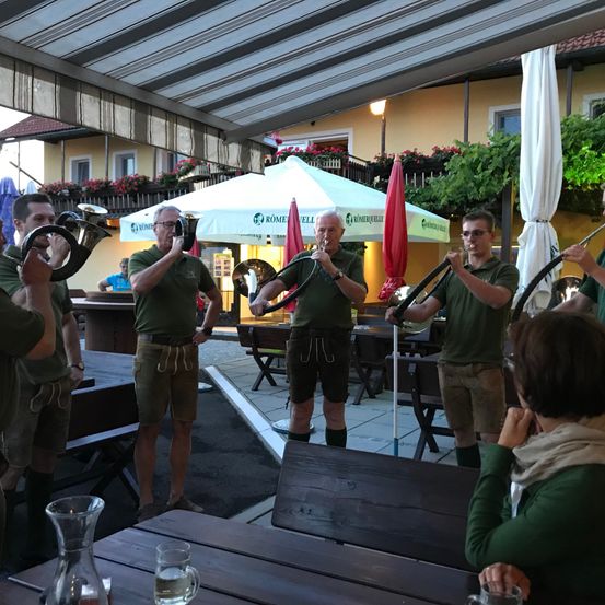Eine Gruppe von Männern in grünen Uniformen spielt Blechblasinstrumente auf einer Terrasse, mit einer Frau, die an einem Tisch im Vordergrund sitzt. Dahinter sind ein Zelt und ein Gebäude mit Pflanzen und Blumen zu sehen.