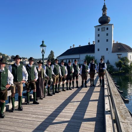 Eine Gruppe von Männern in traditionellen grünen Westen und Hüten steht auf einer Holzbrücke vor einem weißen Gebäude.