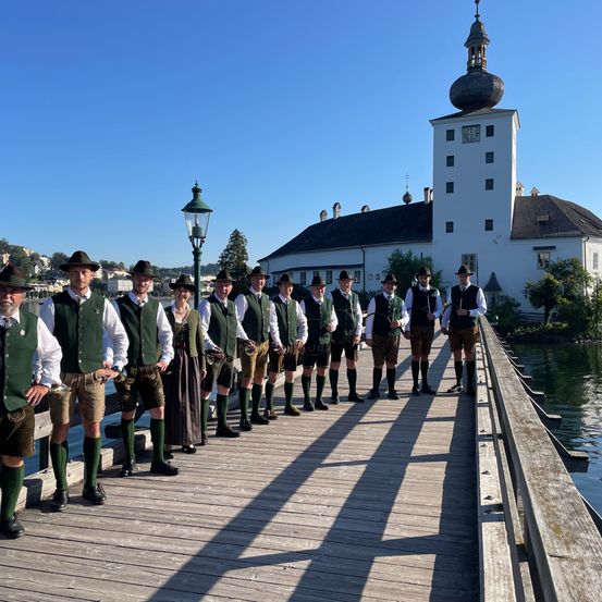 Eine Gruppe von Männern in traditionellen grünen Westen und Hüten steht auf einer Holzbrücke vor einem weißen Gebäude.
