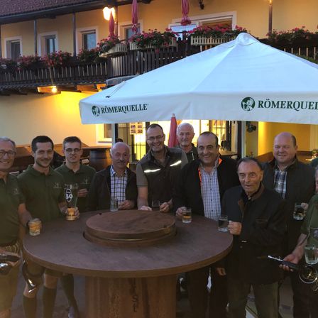 Eine Gruppe von Männern in grünen Shirts steht um einen Holztisch mit Getränken und lächelt für ein Foto. Hinter ihnen steht ein Gebäude mit roten Blumen und ein Schirm mit dem Text 'Römerquelle'.