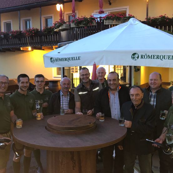 Eine Gruppe von Männern in grünen Shirts steht um einen Holztisch mit Getränken und lächelt für ein Foto. Hinter ihnen steht ein Gebäude mit roten Blumen und ein Schirm mit dem Text 'Römerquelle'.