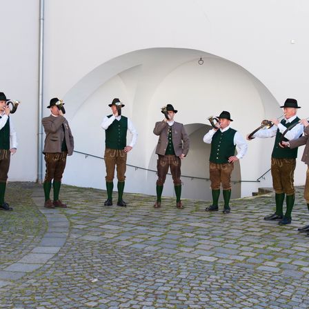 Sechs Männer in traditioneller bayerischer Kleidung, einschließlich grüner Westen und brauner Hosen, stehen in einer Reihe und halten Messinginstrumente. Sie stehen vor einer gewölbten weißen Struktur mit Treppen.