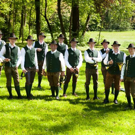 Eine Gruppe von Männern in traditioneller bayerischer Kleidung steht auf einem Rasenfeld und hält Trompeten. Die Männer tragen grüne Westen, braune Hosen und Hüte. Sie sind von Bäumen umgeben.