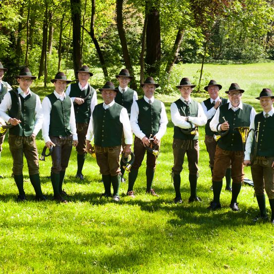 Eine Gruppe von Männern in traditioneller bayerischer Kleidung steht auf einem Rasenfeld und hält Trompeten. Die Männer tragen grüne Westen, braune Hosen und Hüte. Sie sind von Bäumen umgeben.