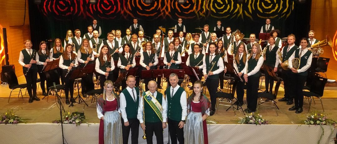 Ein Orchester steht mit verschiedenen Musikinstrumenten auf der Bühne, zwei Männer und zwei Frauen in traditioneller Kleidung davor. Dahinter steht eine größere Gruppe von Orchestermitgliedern mit ihren Instrumenten. Die Bühne ist mit grünen und weißen Blumen und einem bunten Hintergrund geschmückt.