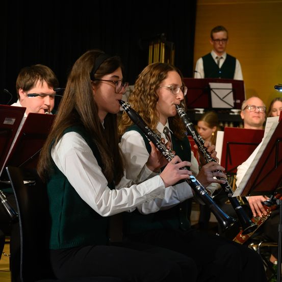 Zwei junge Frauen spielen Klarinette in einer Schulband, sitzend in einem Raum mit anderen Bandmitgliedern und Musikständen im Hintergrund.