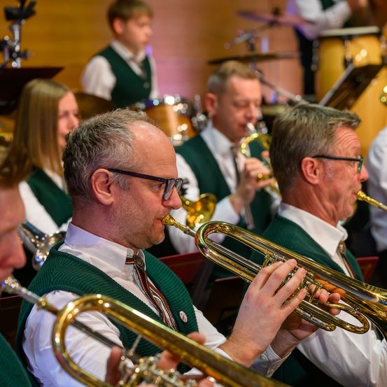 Eine Gruppe von Musikern in grünen Westen und weißen Hemden spielt Blechblasinstrumente bei einer Aufführung. Die Musiker konzentrieren sich auf ihre Instrumente, einer hält eine Trompete und ein anderer eine Posaune. Dahinter spielen weitere Musiker und ein Schlagzeuger.