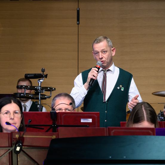 Ein Mann mit einem Mikrofon spricht vor einer Band. Dahinter spielt ein Schlagzeuger. Vorne hört eine Frau mit Brille zu.