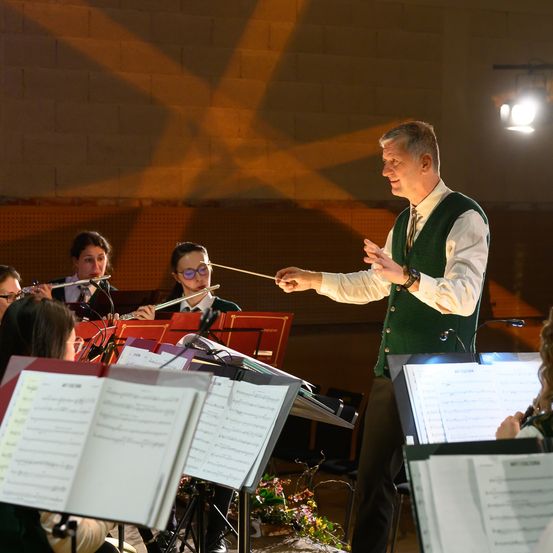 Ein Mann dirigiert ein Orchester mit einem Taktstock, beleuchtet von einer Scheinwerfer. Musiker spielen Flöten und andere Instrumente, mit Notenständern im Vordergrund.