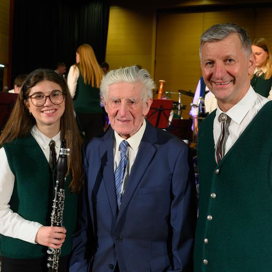 Ein älterer Mann und ein junges Mädchen in einem grünen Weste mit einer Klarinette posieren für ein Foto mit einem weiteren lächelnden Mann. Hinter ihnen ist eine verschwommene Band-Einrichtung.