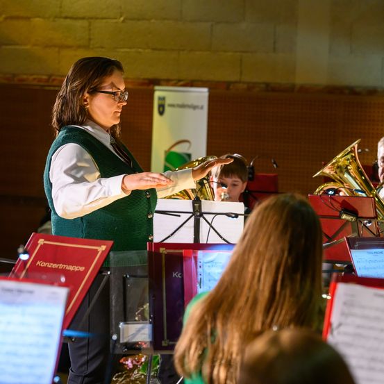 Eine Frau dirigiert ein Orchester mit Notenblättern und Blechblasinstrumenten in einem Raum.