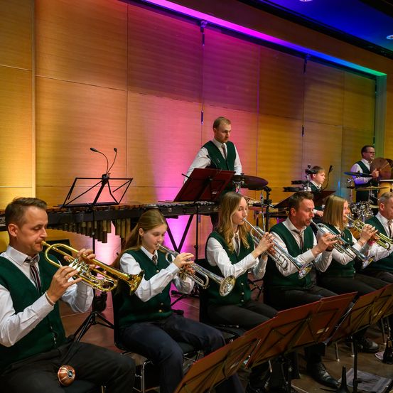 Eine Gruppe von Musikern, gekleidet in grünen Westen, spielen Trompeten in einem Raum mit bunter Beleuchtung. Der Mann im Hintergrund spielt die Trommeln, und einige andere Personen spielen Trommeln und Becken.