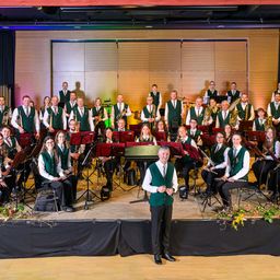 Eine Gruppe Musiker, gekleidet in grünen und weißen Westen, posiert für ein Foto auf einer Bühne mit Musikinstrumenten und einem Dirigenten vor ihnen.