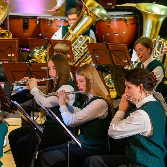 Eine Gruppe von Musikern tritt in einer Indoor-Umgebung auf. Mehrere Frauen sitzen und spielen Flöten, während andere Blechblasinstrumente spielen. Die Musiker sind in grünen Westen und weißen langen Ärmeln gekleidet.
