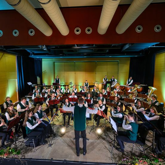 Eine Brassband spielt auf der Bühne, mit Musikern, die verschiedene Instrumente spielen. Der Dirigent steht vorne und leitet das Ensemble. Die Bühne ist beleuchtet, und das Publikum schaut zu.