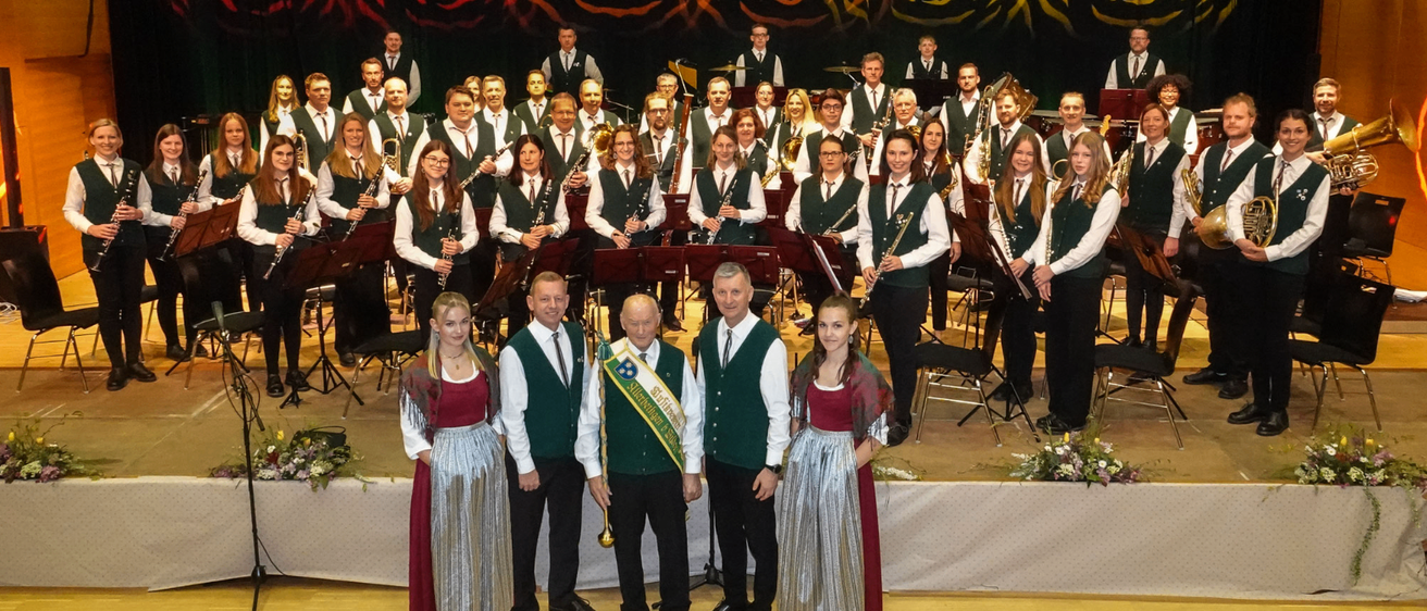 Ein großes Ensemble von Musikern in grünen und weißen Westen und roten Röcken posiert auf der Bühne, wobei einige Instrumente halten. Zwei Männer halten eine Flagge. Dahinter ist die Bühne mit einem farbenfrohen Hintergrund aus Wirbeln dekoriert.