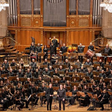 Ein großes Orchester steht auf der Bühne in einem Konzertsaal mit Musikinstrumenten wie Trompeten, Posaunen und Violinen.