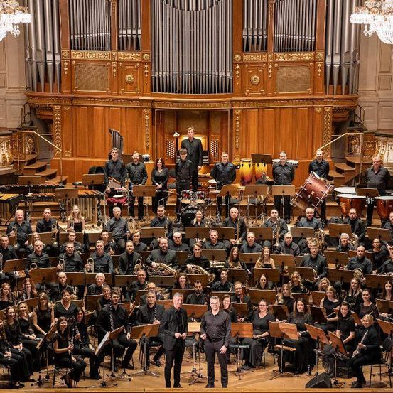 Ein großes Orchester steht auf der Bühne in einem Konzertsaal mit Musikinstrumenten wie Trompeten, Posaunen und Violinen.