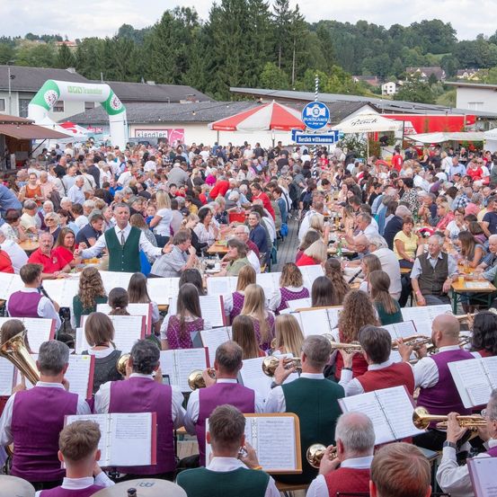 Eine große Menschenmenge ist um Tische herum versammelt, möglicherweise bei einem Open-Air-Festival. Einige tragen traditionelle Kleidung. Eine Band spielt vor ihnen Musik, wobei die Mitglieder verschiedene Instrumente halten.