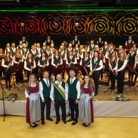 Eine Gruppe von Musikern in grünen Westen und Krawatten posiert auf der Bühne, mit zwei Männern und zwei Frauen davor, und Blumen und Instrumenten im Hintergrund.