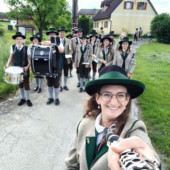 Eine Gruppe von Musikern, einige mit Instrumenten, gehen auf einem Pfad, mit einer lächelnden Frau vor ihnen. Sie tragen alle einheitliche Kleidung.