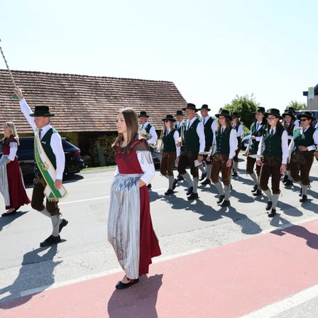 Ein Umzug von Personen in traditioneller Kleidung, die in einer Reihe marschieren, ein Mann hält vorne eine Flagge. Hinter ihm führt eine Frau in einem roten Kleid und Rock die Gruppe an. Die Szene spielt sich draußen ab, mit einem Haus und Grün im Hintergrund.