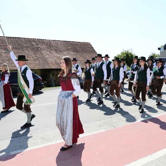 Ein Umzug von Personen in traditioneller Kleidung, die in einer Reihe marschieren, ein Mann hält vorne eine Flagge. Hinter ihm führt eine Frau in einem roten Kleid und Rock die Gruppe an. Die Szene spielt sich draußen ab, mit einem Haus und Grün im Hintergrund.