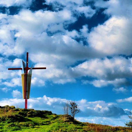 Ein großes Kreuz auf einem grasbewachsenen Hügel unter einem bewölkten Himmel. Das Kreuz hat ein farbenfrohes Design.