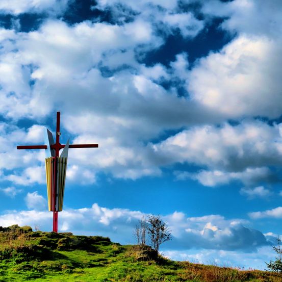 Ein großes Kreuz auf einem grasbewachsenen Hügel unter einem bewölkten Himmel. Das Kreuz hat ein farbenfrohes Design.