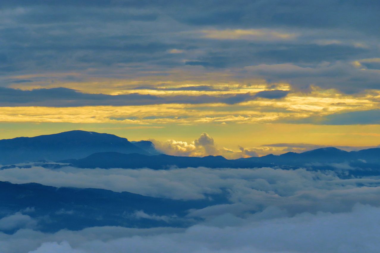Ein atemberaubender Luftblick auf Berge mit einem goldenen Sonnenuntergang, der die Wolken erleuchtet. Der Himmel ist eine Mischung aus Blau und Gelb, und es gibt flauschige Wolken, die über den Gipfeln schweben.