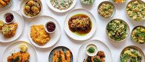 Eine Auswahl an Gerichten ist auf einer weißen Oberfläche angeordnet, einschließlich gebratener Fische, Reis, Salat und andere Lebensmittel.