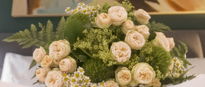 Ein Blumenarrangement mit weißen Rosen, Gänseblümchen und grünen Blättern auf einem weißen Tisch, mit einem farbenfrohen Gemälde im Hintergrund.