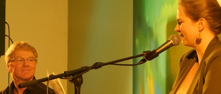 A woman is singing into a microphone while playing the piano, with a man sitting and smiling next to her.