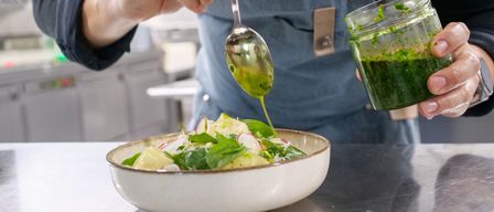 Ein Koch gießt eine grüne Sauce aus einem Glas über einen Salat in einer Schüssel. Der Koch trägt eine Schürze mit Logo.