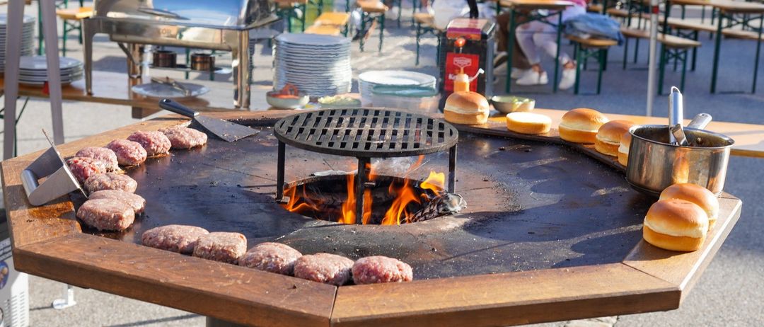 Ein großer Outdoor-Grill mit rohen Hamburgern, umgeben von einer Gruppe von Menschen, die an Tischen in einem Biergarten sitzen.