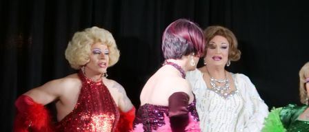Three women in glamorous attire, one with blonde wig and red dress, another with pink hair and sequined dress, and the third in a white dress with elaborate makeup.